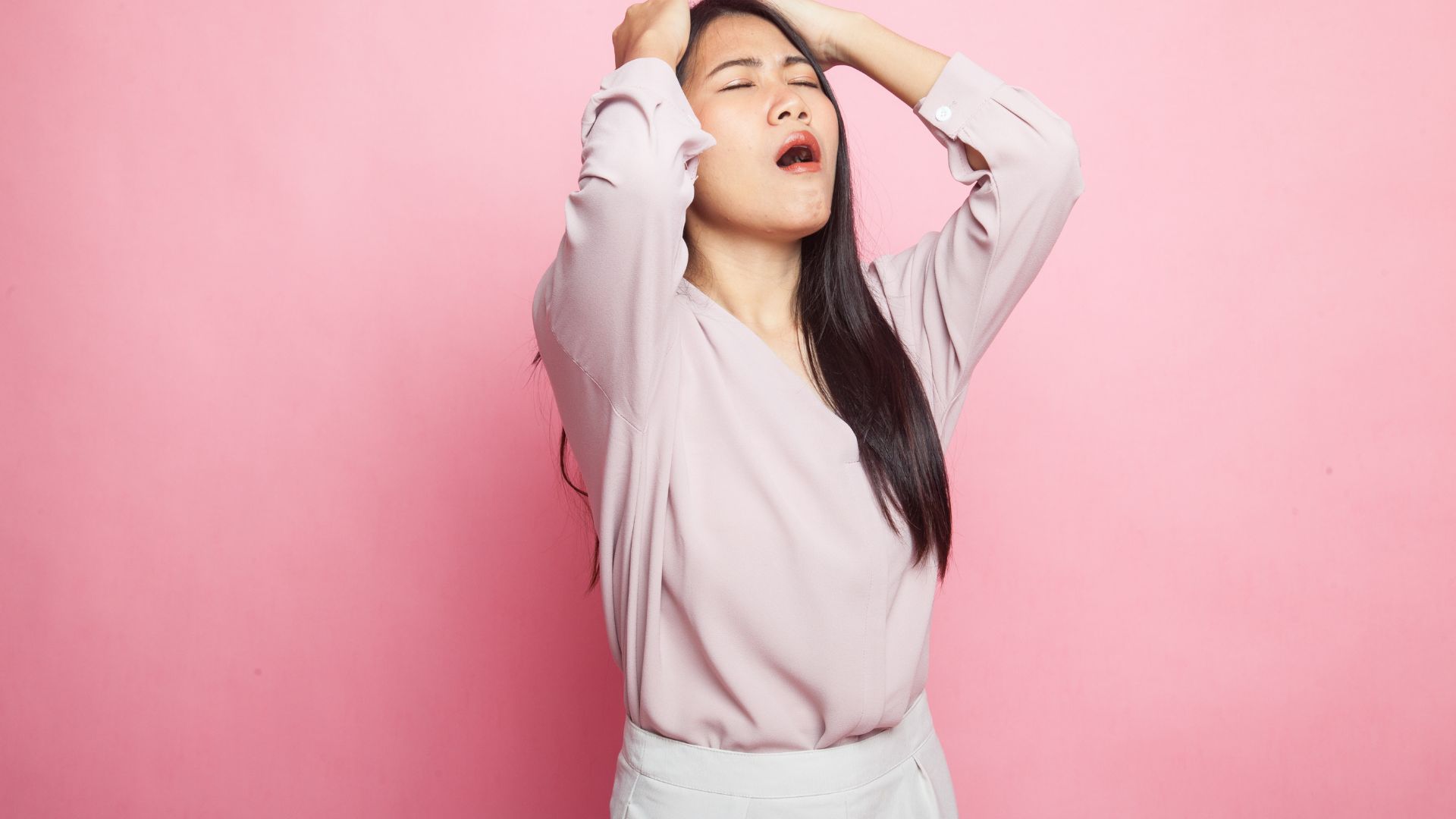 ピンクの背景で頭を抱え、ストレスによる精神的な苦痛を表現するアジア人女性。心身の不調は専門家によるケアで改善が期待できます。